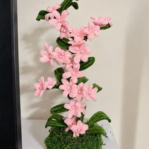 Pode incluir: Uma planta decorativa com flores cor-de-rosa claro de cinco pétalas feitas de material aveludado, cada uma com uma pequena pérola branca no centro. As flores estão presas a caules verdes texturizados com folhas, sobre uma base de musgo.