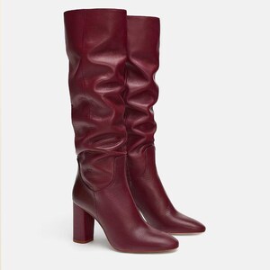May include: A pair of knee-high boots in a deep burgundy color. These boots feature a slouchy, gathered shaft design, a rounded toe, and a sturdy block heel. The exterior has a textured finish, and the boots reach up to the lower calf area.