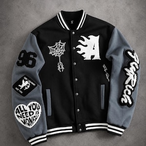 Op de afbeelding: Een zwart en grijs varsity-jack met witte geborduurde patches, waaronder een spinnenweb, de letter A en teksten zoals 96, Don't Be Afraid Not You, All You Need Is Money en Fly Rich. Het jack heeft een drukknoopsluiting en gestreepte boorden.