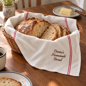May include: A woven wicker basket lined with a white cotton towel featuring two vertical red stripes and the embroidered text "Denise's Homemade Bread" in brown, containing several slices of crusty, golden-brown sourdough bread.