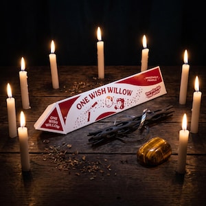 May include: A triangular white and red box labeled ONE WISH WILLOW with illustrations of two faces, a dark brown textured object, and a smooth golden-brown stone, all surrounded by nine lit white taper candles on a dark wooden surface.