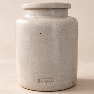 May include: A cylindrical ceramic jar with a light beige, speckled, and textured glaze. The jar features a wide, flat rim and a short neck. The word "Lever" is embossed in a small, rectangular recessed area near the base of the vessel.