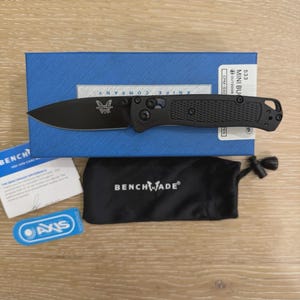 May include: A black folding knife with a textured handle and a drop-point blade featuring the Benchmade logo. It is displayed with a blue box labeled 533 MINI BUG, a black drawstring pouch with the Benchmade logo, and a small blue AXIS tag.