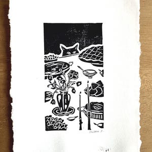 May include: A black and white linocut print on textured paper featuring a cat peeking over a table set with pies, a vase of flowers, and candles. The artwork depicts a dining scene with various dishes and is signed in the bottom right corner as Shima S.