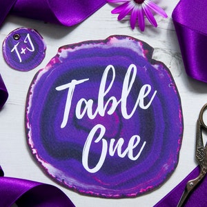 May include: Purple agate table number card with white text that reads "Table One". A small purple and black circle tag with the initials "T+J" is also visible.