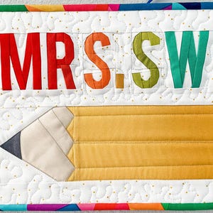 May include: A white quilted fabric banner with the text "MRS. SWA" in colorful fabric letters. A yellow and white pencil is sewn on the bottom of the banner.