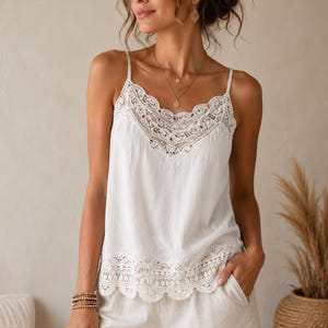 Puede incluir: Una camiseta de tirantes blanca sin mangas con un delicado borde de encaje a lo largo del escote en V y el dobladillo inferior. La prenda está confeccionada con un tejido texturizado e incluye tirantes finos, ofreciendo un diseño simple.