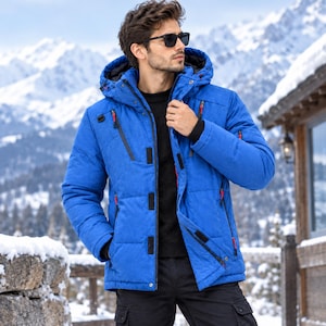 Op de afbeelding: Een felblauwe winterjas met capuchon, zwarte voering, ritssluiting aan de voorzijde met zwarte klittenbandsluitingen en meerdere zakken met kleine rode ritstrekkers, gedragen over een zwart shirt en een zwarte broek.