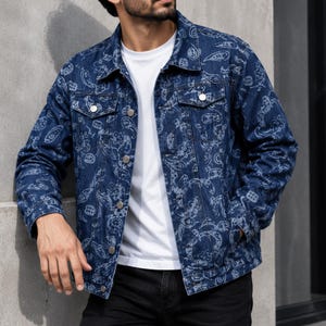 Könnte beinhalten: Eine dunkelblaue Jeansjacke mit einem durchgehenden weißen Paisleymuster. Die Jacke hat einen klassischen Kragen, lange Ärmel, zwei Brusttaschen mit Knopfverschluss und eine Knopfleiste vorne. Sie wird über einem schlichten weißen T-Shirt getragen.