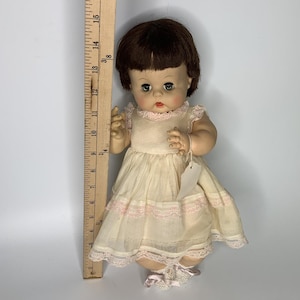 Könnte beinhalten: Eine Puppe mit braunen Haaren und grünen Augen trägt ein cremefarbenes Kleid mit rosa Spitzenbesatz. Die Puppe steht neben einem Holzlineal, das eine Höhe von etwa 38 cm anzeigt. Die Puppe trägt passende cremefarbene Schuhe mit rosa Schleifen.