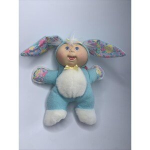 May include: A plush doll dressed in a light blue bunny costume with a white fuzzy belly and feet. The long ears feature a colorful floral pattern in pink, yellow, and blue. The doll has a round face, blue eyes, and a small yellow bow at the neck.