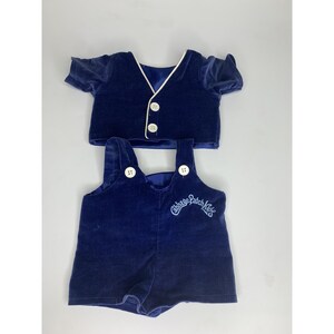 May include: A dark blue velvet doll outfit consisting of a short-sleeved vest with white trim and two white buttons, and matching overalls with two white buttons on the straps. The overalls feature the embroidered text Cabbage Patch Kids.