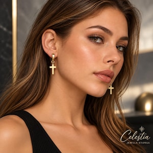 May include: A pair of gold-toned hoop earrings featuring a dangling cross charm. The earrings have a polished finish and a secure hinged closure. The text Celestia JEWELS STONES is visible in the bottom right corner of the image.