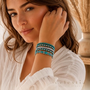 Op de afbeelding: Een armband met meerdere strengen, voorzien van turkooiskleurige ovale stenen in zilverkleurig metaal, afgewisseld met kleine zilverkleurige kralen en een decoratieve zilverkleurige metalen band in filigraanstijl, gedragen bij een wit gestructureerd topje.