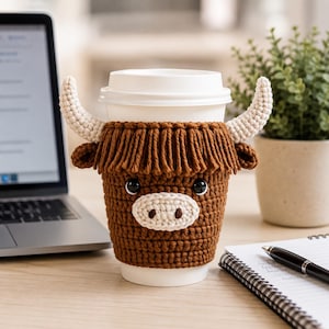 May include: A brown crocheted coffee cup sleeve designed to look like a Highland cow, featuring a textured fringe, two cream-colored horns, small ears, black button eyes, and a cream-colored snout with two dark brown nostrils on a white cup.