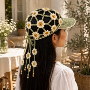 May include: A crochet baseball cap featuring a sage green lattice pattern adorned with white and yellow daisy motifs. The cap includes a solid sage green brim and long, dangling crochet ties at the back, each finished with small daisy accents and beads.