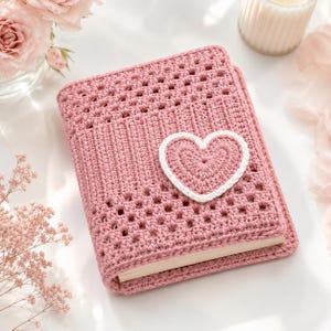 Può includere: Una copertina per libro rettangolare realizzata in filato rosa polvere, caratterizzata da un motivo a uncinetto testurizzato con un design a griglia. Un cuore rosa polvere pieno con un bordo spesso a uncinetto bianco è centrato sulla copertina.