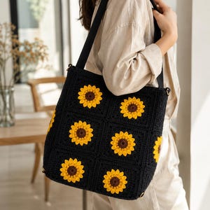 May include: A black crochet tote bag featuring a pattern of square motifs, each centered with a vibrant yellow and brown sunflower design. The bag includes a solid black fabric shoulder strap attached to the top corners with gold-toned metal hardware.