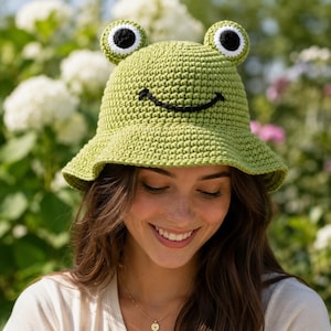 May include: A light green crochet bucket hat featuring two protruding frog eyes with white and black centres and a stitched black curved mouth on the front. The hat is constructed with a textured yarn stitch pattern and a wide, downward-sloping brim.