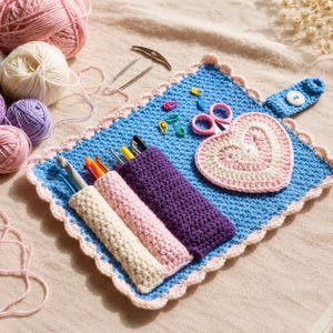 Pode incluir: Um estojo organizador de crochê azul com borda festonada rosa claro, contendo três bolsos com agulhas de crochê, uma aplicação em forma de coração rosa, tesoura pequena e marcadores de pontos coloridos. Um botão branco fecha a aba.