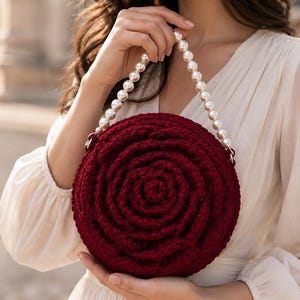 May include: A round, dark red crochet handbag featuring a textured, spiraling floral pattern. The bag is equipped with a single handle made of a string of white, spherical faux pearls attached to the top sides of the circular, yarn-based structure.