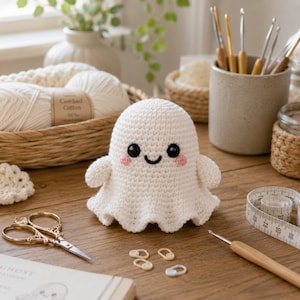 Könnte beinhalten: Ein weißes, gehäkeltes Geister-Amigurumi mit schwarzen Sicherheitsaugen und kleinen rosa Wangen. Der Geist ist umgeben von Bastelbedarf, darunter ein Korb mit weißem Garn, ein Keramikbecher mit Häkelnadeln, eine goldene Schere und ein Maßband.
