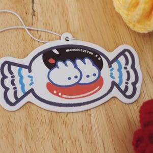 May include: A white, candy-shaped air freshener featuring a graphic illustration of two small white bunnies with black eyes inside a dark opening, a red mouth, and blue accents on the wrapper ends. The text "CODEDCOZY" is printed in small white letters.