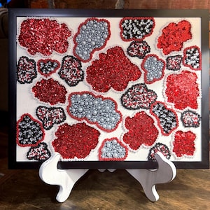 May include: A framed wall art piece featuring an abstract arrangement of irregular shapes filled with red glitter, silver pearls, and black and clear rhinestones on a white background, all outlined with small white beads in a black frame.