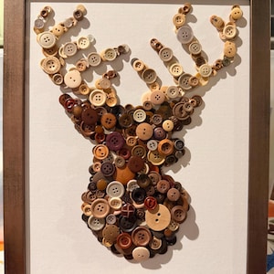 Puede incluir: Una pieza de arte mural enmarcada que presenta la silueta de una cabeza de ciervo construida con numerosos botones redondos en varios tonos de marrón, bronceado, crema y gris. Los botones están dispuestos sobre un fondo blanco en un marco de color bronce.
