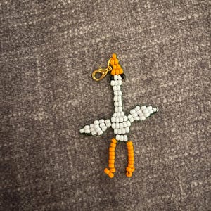 May include: A beaded bird charm featuring a white body and wings, an orange head, and orange legs. The bird is constructed from small, uniform beads and includes a gold-toned metal lobster claw clasp attached to the top of the head for hanging.