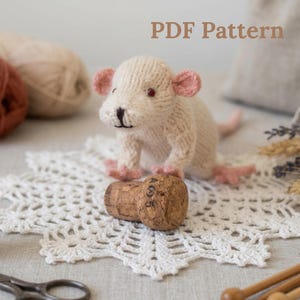 May include: A hand-knitted cream-colored mouse toy with pink inner ears and feet, featuring dark eyes and a stitched nose. The mouse rests its front paws on a cylindrical cork stopper marked with the text A05. The image includes the text PDF Pattern.