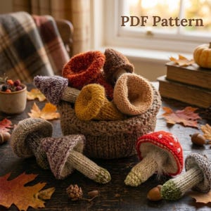 May include: A collection of knitted mushroom figures in various colors including red, orange, brown, and purple, arranged in a brown crocheted basket. The text PDF Pattern is visible in the upper right corner. The mushrooms feature textured caps and stems.