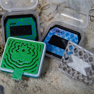 May include: Two clear plastic cases containing square ink pads with blue and green patterned surfaces labeled ARAT. A bright green square stamp with a black topographic line design and a silver geometric lattice stamp are positioned in front.