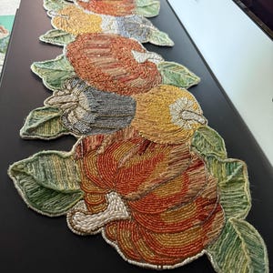 May include: A beaded table runner featuring a repeating pattern of pumpkins and gourds in shades of orange, yellow, and grey, accented with green leaves. The entire surface is covered in intricate, small glass beads arranged in rows.