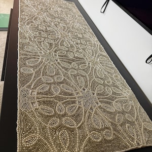 May include: A rectangular table runner featuring an intricate, repeating floral pattern composed of small, shimmering silver and white beads. The edges are finished with a border of tiny white beads, creating a textured, metallic surface.