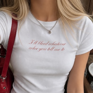 Może przedstawiać: Biały t-shirt z krótkim rękawem i okrągłym dekoltem, z czerwonym napisem "I'll bleed whatever color you tell me to" na klatce piersiowej. Koszulka zestawiona jest z niebieskimi dżinsami, czerwoną torebką na ramię oraz srebrnym naszyjnikiem z sercem.