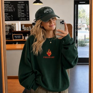 Puede incluir: Una sudadera de cuello redondo de color verde oscuro con un pequeño gráfico bordado de un corazón rojo rodeado de llamas naranjas y amarillas, con la palabra FIREHEART impresa en rojo debajo. También se ve una gorra de béisbol verde oscuro con texto blanco.
