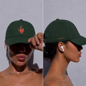 Puede incluir: Una gorra de béisbol de color verde oscuro con un corazón rojo y naranja bordado rodeado de llamas en el panel frontal. El lateral de la gorra muestra el texto FIREHEART en letras pequeñas de color naranja e incluye una correa metálica ajustable.