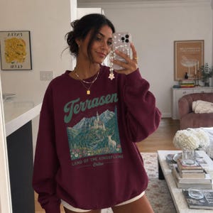May include: A maroon long-sleeve crewneck sweatshirt featuring a graphic print of a mountain landscape, a castle, a white deer, and colourful flowers. The text reads Terrasen, Land of the Kingsflame, and Erilea in a mix of script and block fonts.