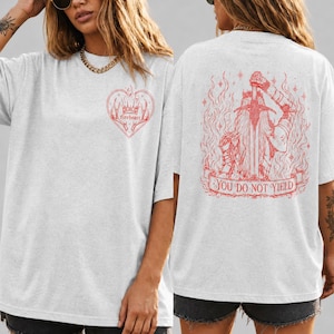 Puede incluir: Una camiseta gris claro con un estampado gráfico rojo. La parte delantera muestra un corazón con una corona y el texto fireheart. La parte trasera muestra una figura sosteniendo una espada rodeada de llamas con una cinta con el texto YOU DO NOT YIELD.