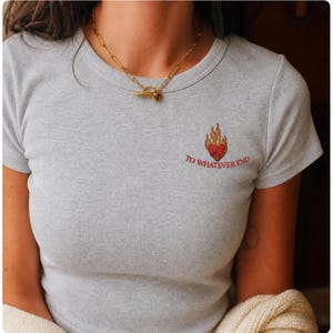 May include: A light grey ribbed short-sleeve t-shirt featuring an embroidered design on the left chest. The embroidery depicts a red heart surrounded by orange and yellow flames, with the text TO WHATEVER END stitched in red underneath.