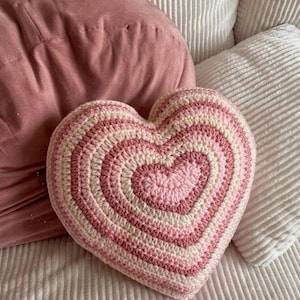Puede incluir: Un cojín con forma de corazón hecho de ganchillo con anillos concéntricos de hilo en tonos alternos de rosa claro, rosa oscuro y crema. La superficie texturizada muestra un patrón de puntos consistente en toda la forma del corazón.