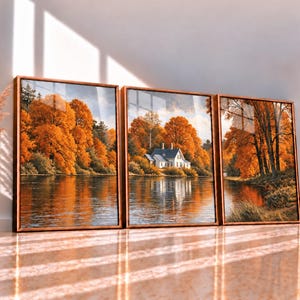 May include: A triptych wall art set featuring a landscape painting of a white house nestled among vibrant orange autumn trees beside a calm river. The three framed prints are displayed side-by-side, accompanied by a tall vase with dried grass.