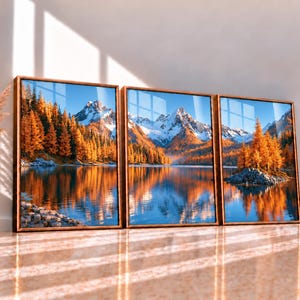 May include: A triptych wall art set featuring a landscape of snow-capped mountains, a calm lake, and golden autumn trees, all framed in wood. Beside the art stands a tall, light-colored ceramic vase filled with dried pampas grass.