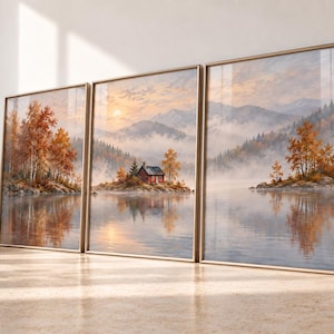 May include: A triptych wall art set featuring a landscape painting of a small red cabin on a misty island in a lake, surrounded by autumn trees and mountains at sunrise. The three framed prints are displayed side-by-side in thin gold frames.