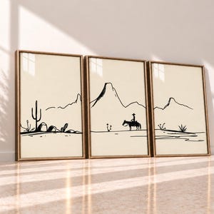 May include: A set of three framed wall art prints featuring black line drawings on a cream background. The illustrations depict a desert landscape with mountains, a saguaro cactus, rocks, and a person riding a horse, all enclosed in wooden frames.