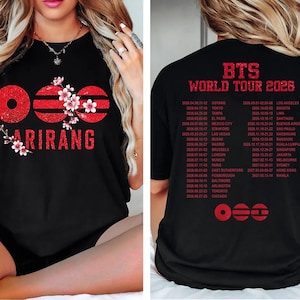 Op de afbeelding: Een zwart t-shirt met een rode glitterafbeelding op de voorkant met de tekst ARIRANG en kersenbloesemaccenten. De achterkant toont de tekst BTS WORLD TOUR 2026 in het rood, gevolgd door een lijst met tourdata en steden in twee kolommen.