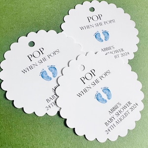 Może przedstawiać: Trzy białe, faliste, okrągłe etykiety z niebieskimi śladami stóp dziecka i tekstem "POP WHEN SHE POPS!" Etykiety są na baby shower i zawierają imię Abbie's oraz datę 24 sierpnia 2024.