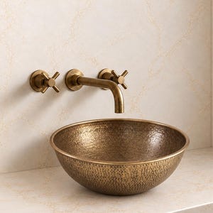 May include: A round, hammered bronze vessel sink with a textured finish. Above the sink, a wall-mounted bronze faucet features a curved spout and two cross-handle valves, all finished in a matching metallic tone to coordinate with the basin.