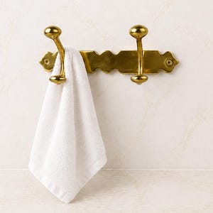May include: A polished brass wall-mounted coat rack featuring two double-pronged hooks with rounded finials, mounted on a decorative rectangular backplate with scalloped edges, holding a single plain white folded towel on the left hook.